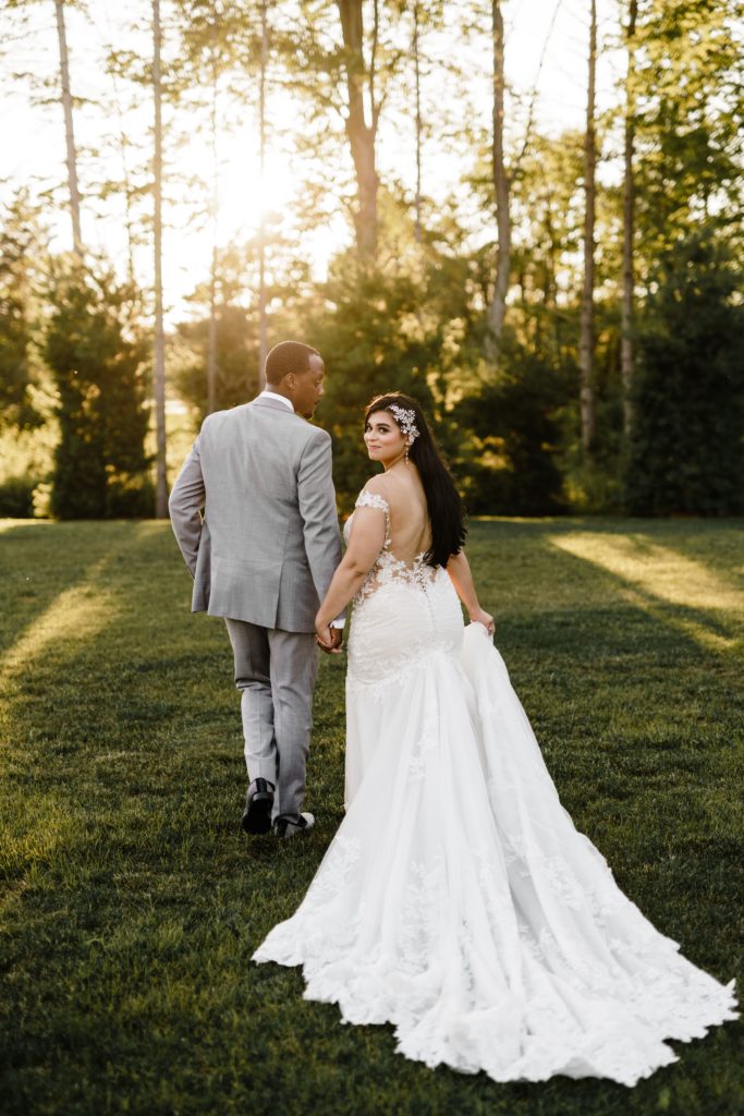 the bride looks over her shoulder as the groom holding her hand walks across an open field toward the shining golden sun as their luxury wedding photographer trails to get this photo
