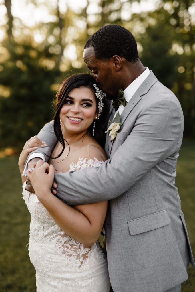 the brides smiles as the groom presses a kiss to the crown of her head and wraps his arms around her shoulders as they pose for luxury wedding photography