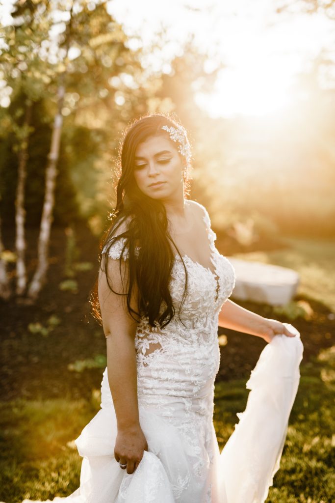 the bride looks down while holding her dress while posing for timeless wedding photography with golden light streaming behind her