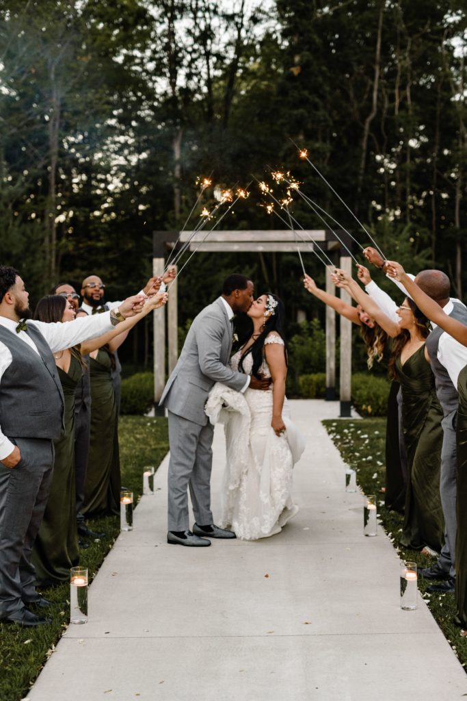 the bride and groom kiss under a sparkler tunnel held up by their bridal party in front of their wedding arch while luxury wedding photographer captures the moment
