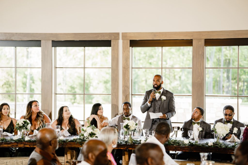 the best man talks into a microphone during his speech at the black river barn wedding