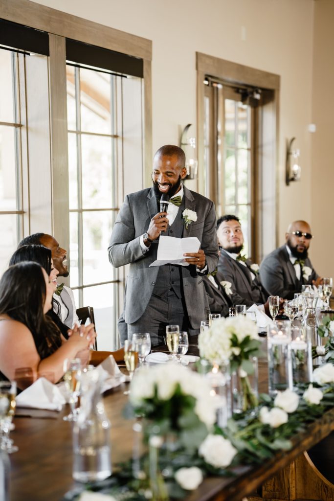 the best man laughs with the groom as he gives a speech while standing behind the head table captured by the michigan wedding photographer