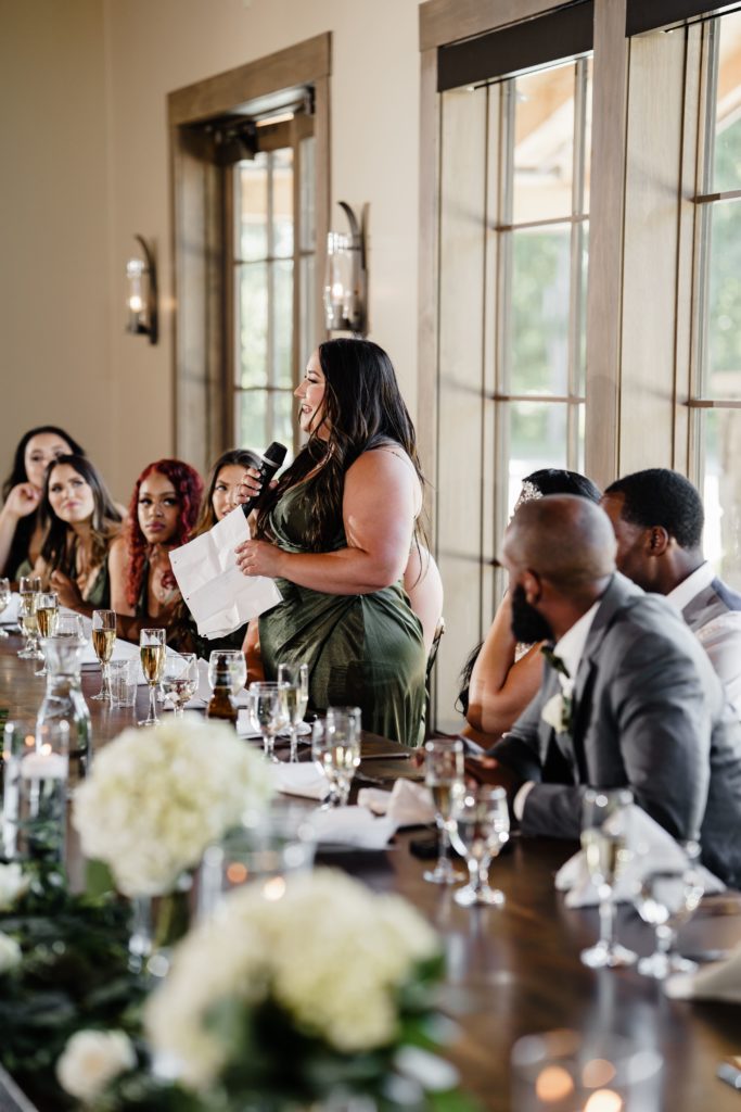 the maid of honor stands holding a microphone and a sheet of paper at her place at the head table while giving a speech as the couple's luxury wedding photographer captures the day