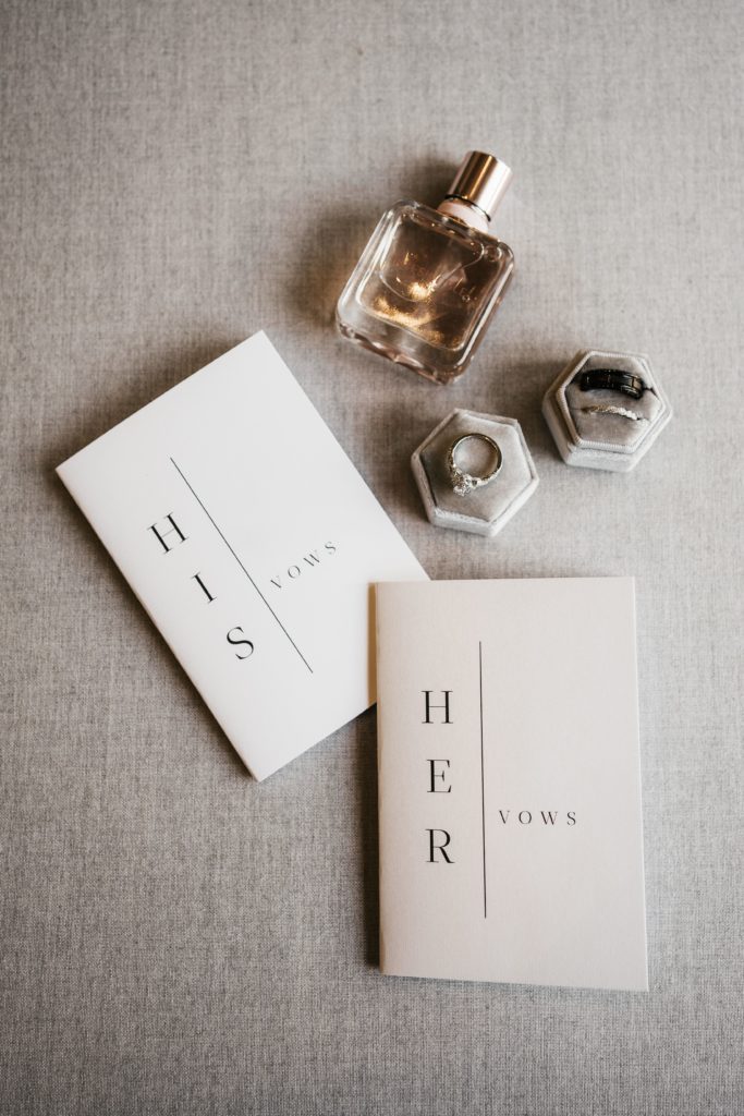 a detail shot from a michigan wedding, vow books lay next to rings resting in boxes next to the bride's perfume taken by a michigan wedding photographer