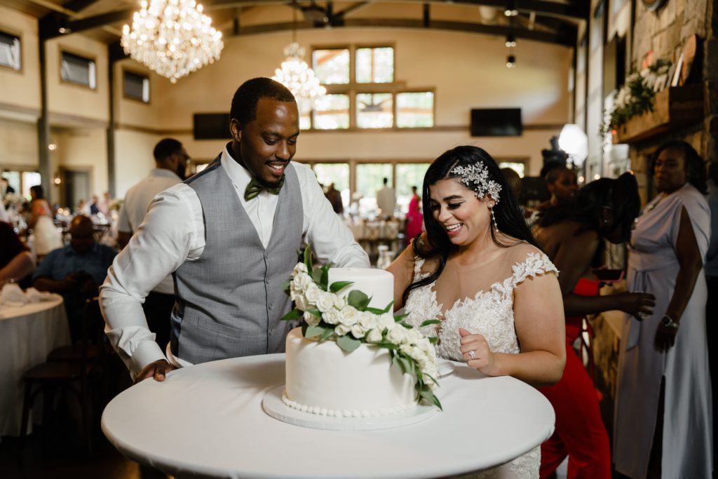 the bride and groom smile as they cut into their classic two tier wedding cake while being captured by their luxury wedding photographer