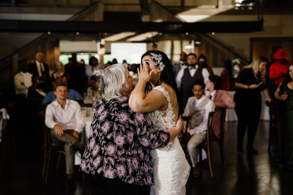 the bride wipes away a tear as she shares a dance with her mom while remembering her father who passed. her michigan wedding photographer uses the light of the reception hall to help convey the way the moment feels