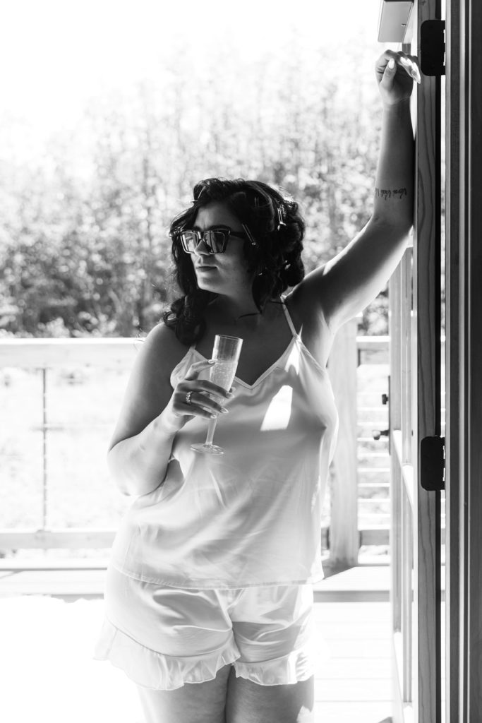 the bride leans against a door while holding a glass of champagne, wearing sunglasses and silk pajamas, and prepping her hair as michigan wedding photographer documents the day