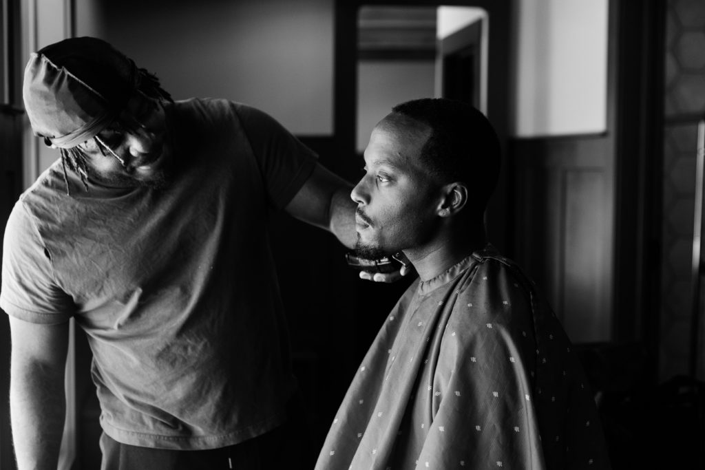 the groom shares a special moment with his friend who is a barber as he gives him a fresh trim for his wedding day. this luxury wedding photographer uses lighting to convey the emotion of the moment and the excitement of the day