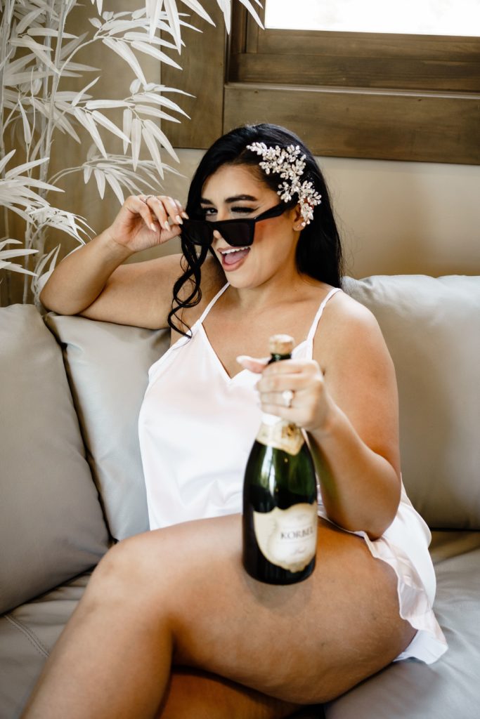 the bride smiles and winks as she holds a bottle of champagne before getting ready for her wedding day as it is documented by a luxury wedding photographer