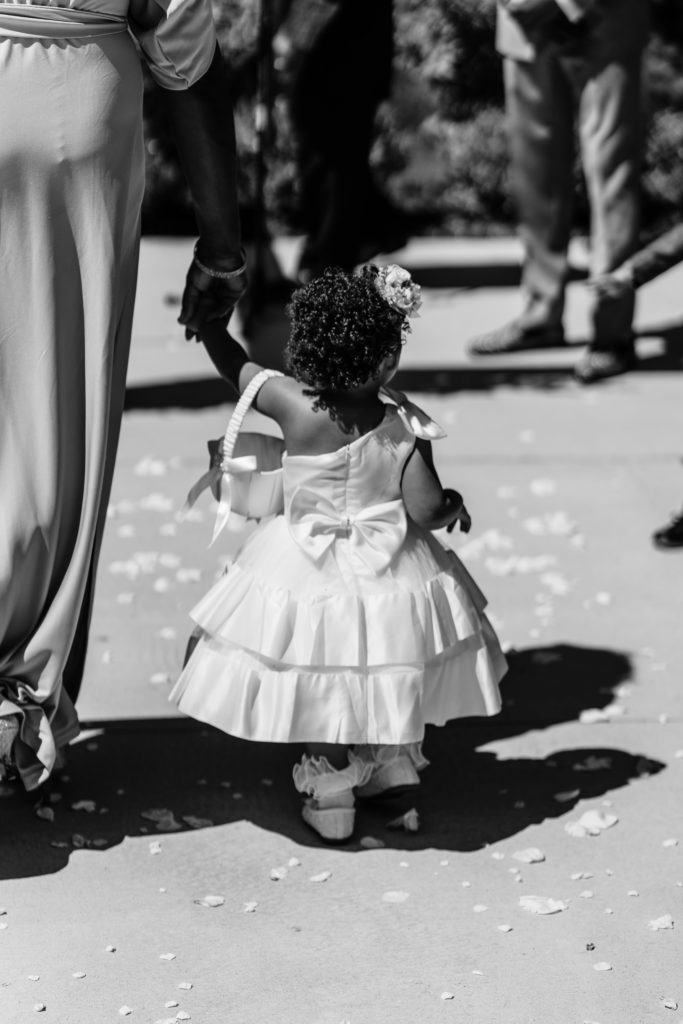 a black and white portrait of the flower girl in a white dress holding an adult's hand as they walk down the aisle as timeless wedding photography is captured