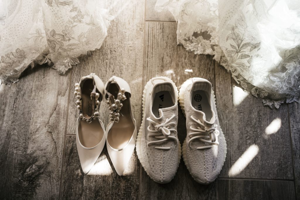 the bride's two pairs of shoes, one pair of heels and one pair of white sneakers, wait while the details of her wedding dress peek out behind them, set out for luxury wedding photography