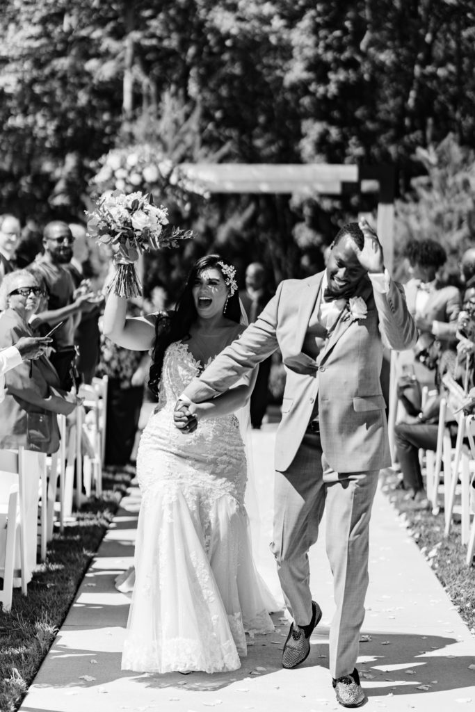 the groom waves as the bride raises her bouquet in celebration as they walk down the aisle together after their wedding ceremony while their photographer snaps their timeless wedding photography
