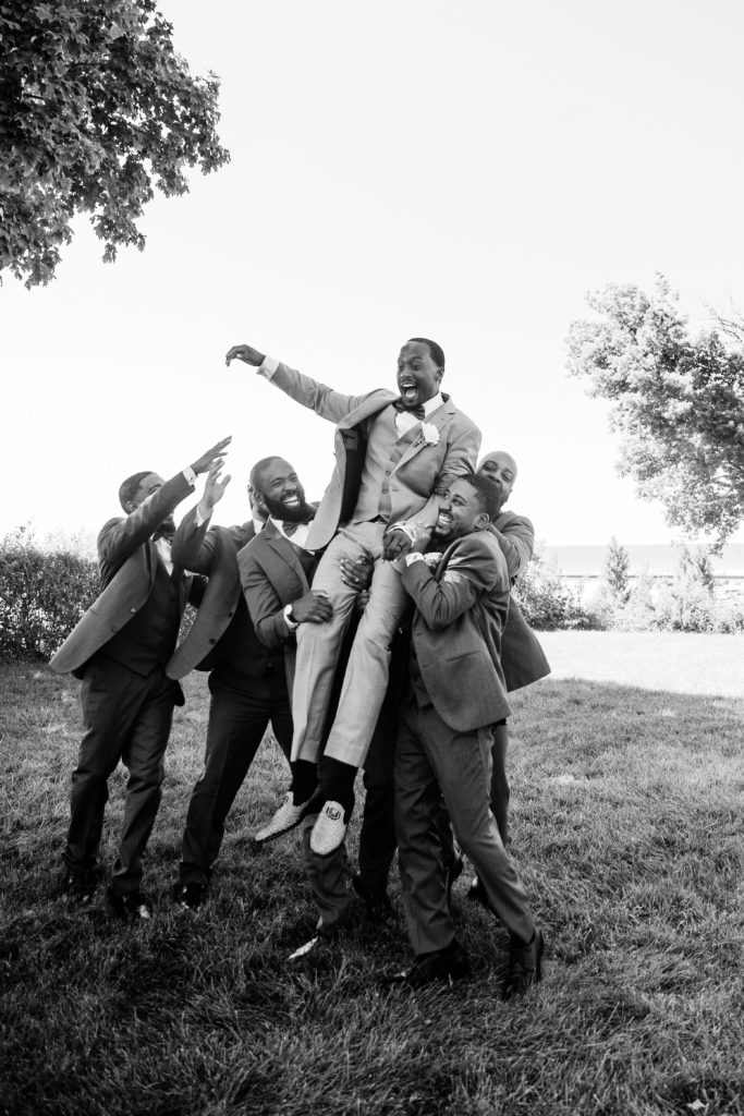 the groomsmen laugh as they lift up the groom together while posing for the michigan wedding photographer