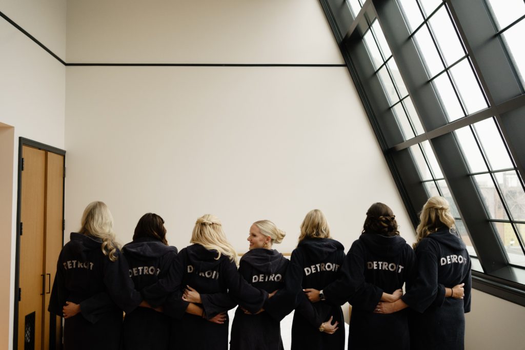 the bride and bridesmaids stand in matching robes that say detroit on the back while getting ready for the shinola hotel wedding