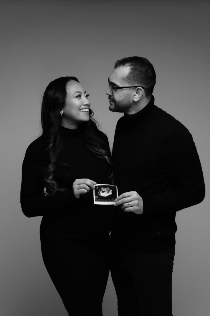 an expecting couple smile at each other while holding their sonogram during their indoor maternity shoot