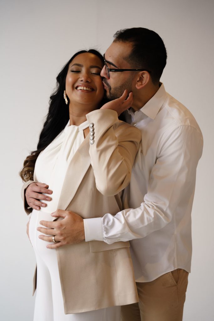 an expecting couple stand in front of a white backdrop with their hands on her belly as he presses a kiss to her cheek during their editorial maternity shoot