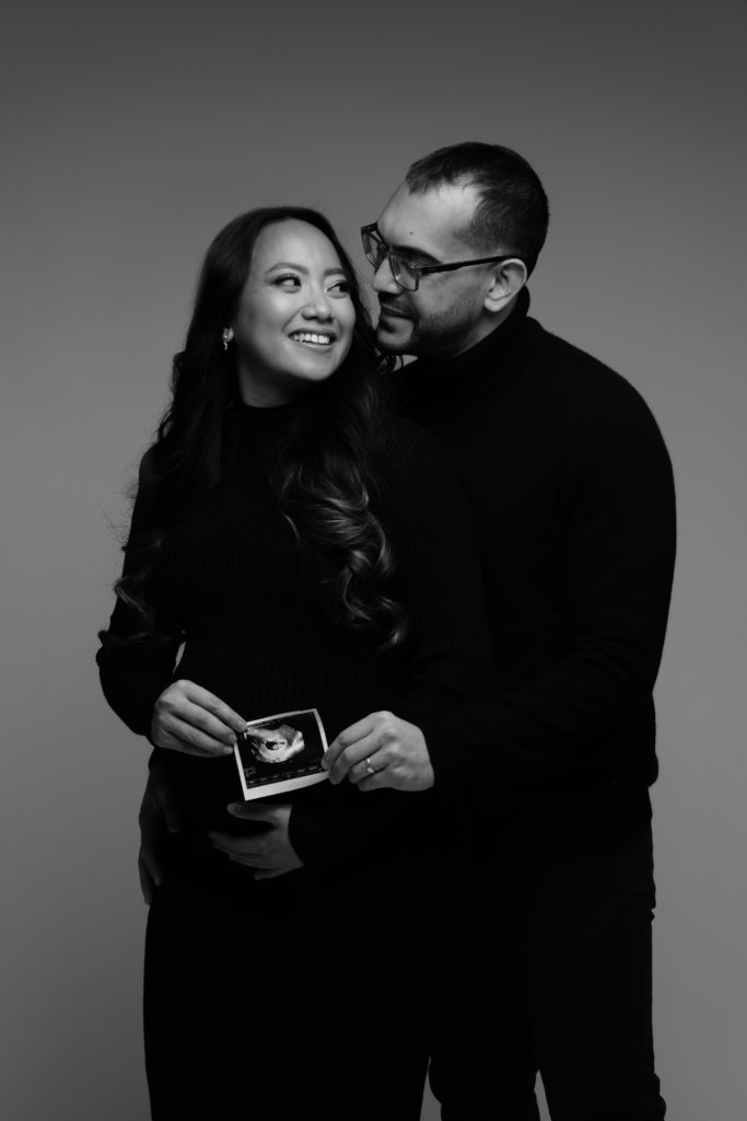 an expecting couple stand smiling at each other while holding their sonogram in front of her belly during their indoor maternity shoot