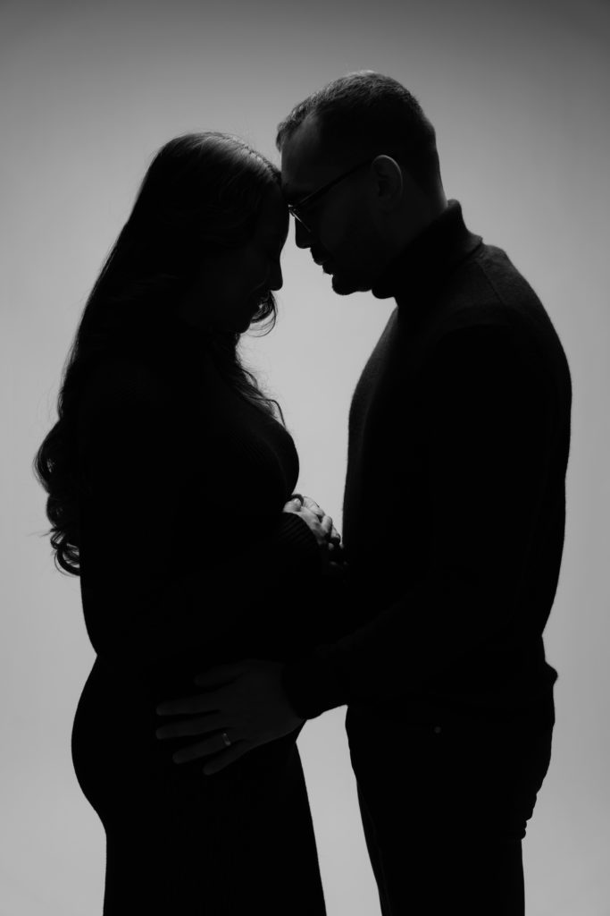 an expectant couple lean on each other while backlit during their luxury maternity photoshoot
