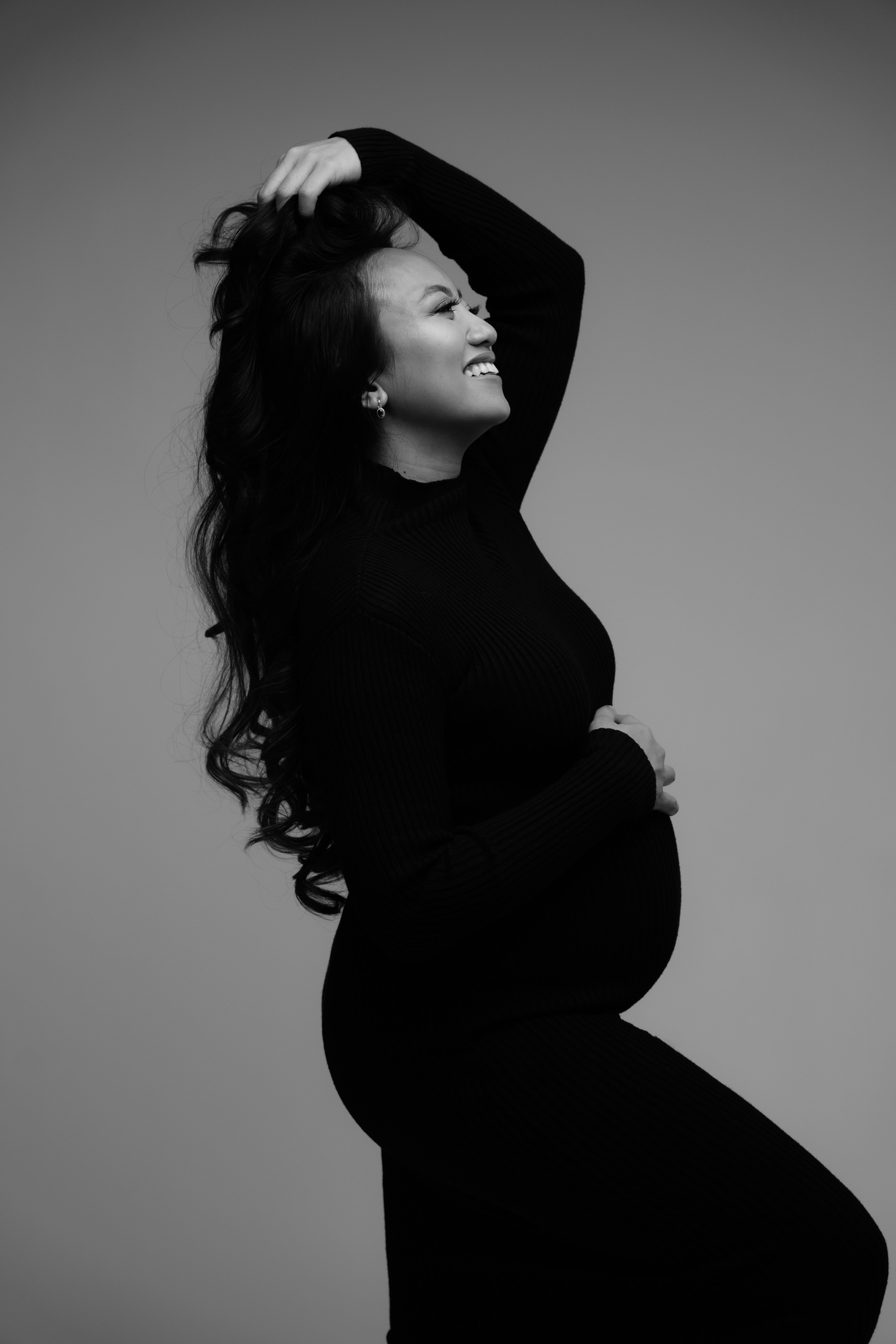 a woman laughs with one hand on her pregnant belly and the other in her hair during her indoor maternity shoot
