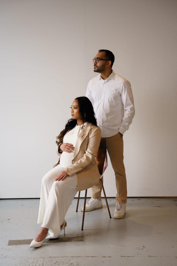an expectant mother sits in a chair with her partner standing behind her and they look off into the distance during their editorial maternity shoot