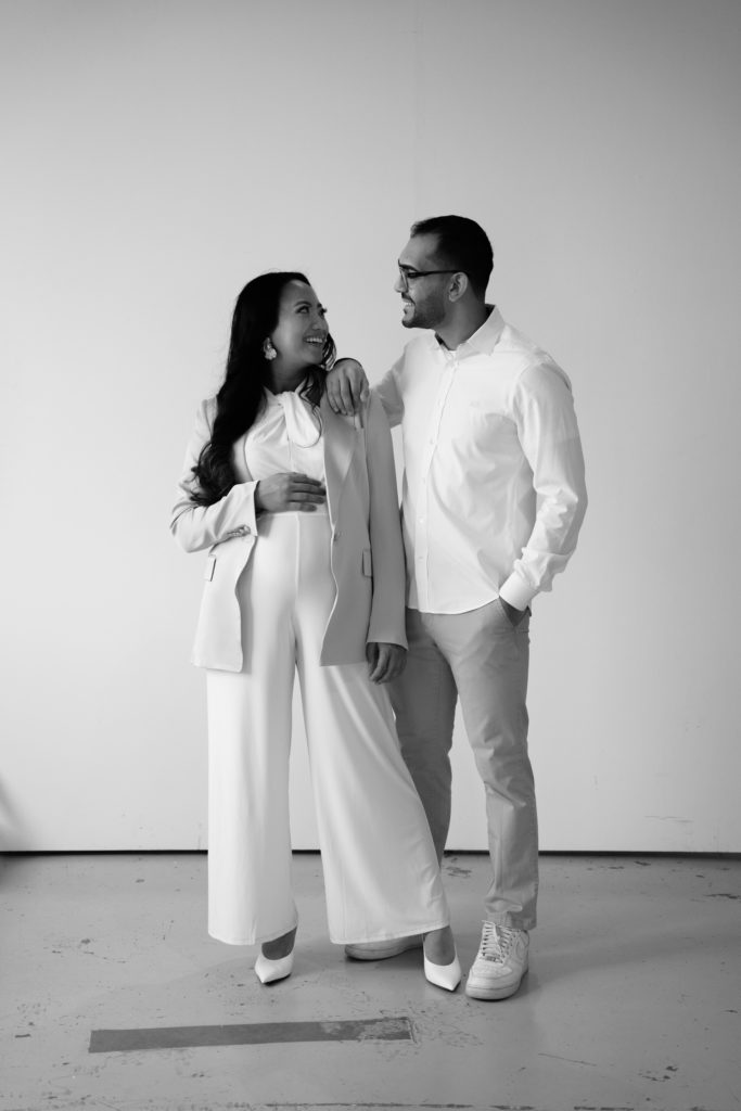 an expecting couple stand smiling at each other during their indoor maternity shoot