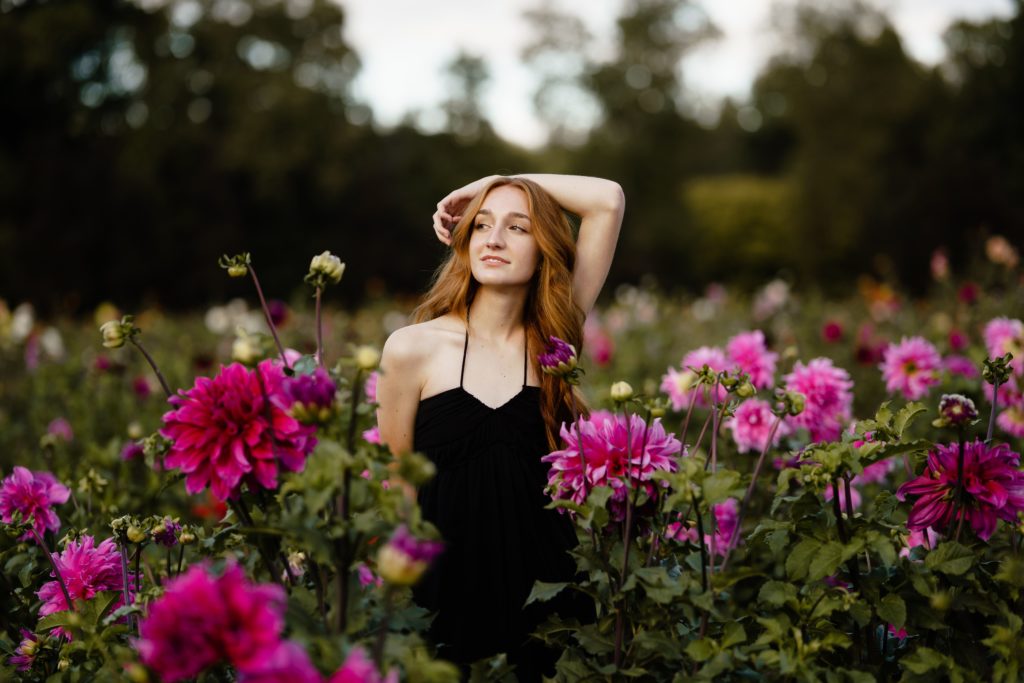 louise stands in a field of flowers with one arm resting over her head while posing for senior portrait photography