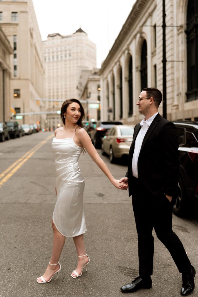 julia looks back at her fiancee as they walk across the downtown detroit street during their engagement photoshoot