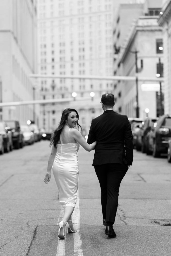 julia looks back over her shoulder as she walks down the street with her fiancee during their detroit engagement photos