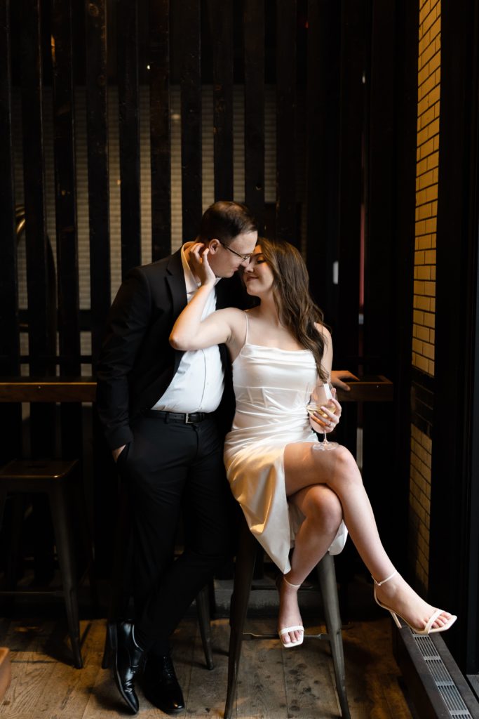 julia sits in a stool as drew leans in towards her during their engagement photoshoot
