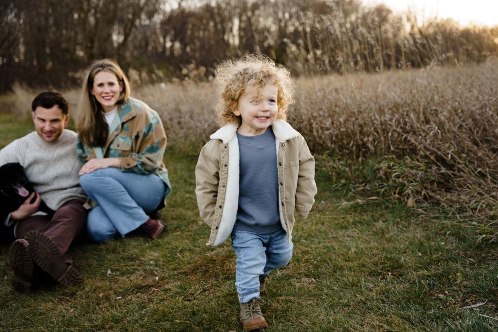 a boy smiles while running in a field with his family in the background during family portraits photography