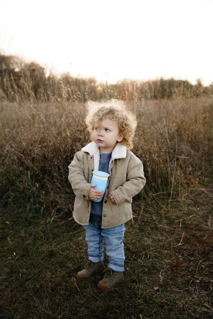 a toddler stands in front of a field clutching his sippy cup during family portraits