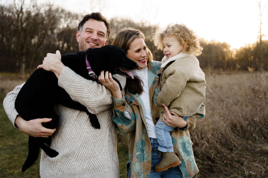 a man holds up the family dog while standing next to his wife who carries their son during family portraits photography