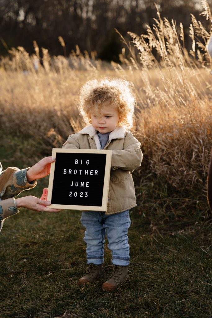 a boy looks down grumpily as he stands in a field while someone holds out a letter board that spells out "big brother june 2023" during their session with a family photographer
