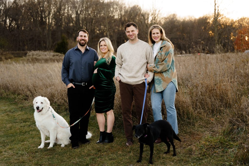 two couples stand in an open field at golden hour with their smiling dogs