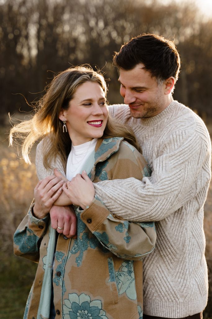 a couple stand with his arms around her shoulders as she looks back at him during family portraits