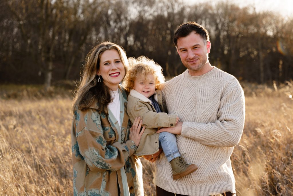 a family of three stand in a field at golden hour smiling during a session with detroit family photographer at maybury state park in northville michigan