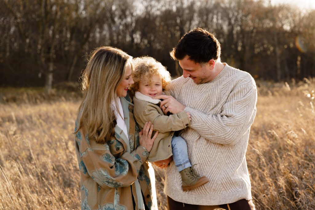 a family of three stand together in a field, dad tickling the toddler during a session with a family photographer