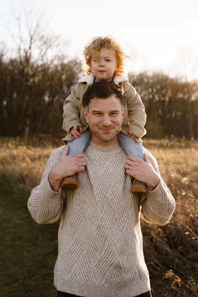 a boy sits on his father's shoulders as they smile at the family photographer