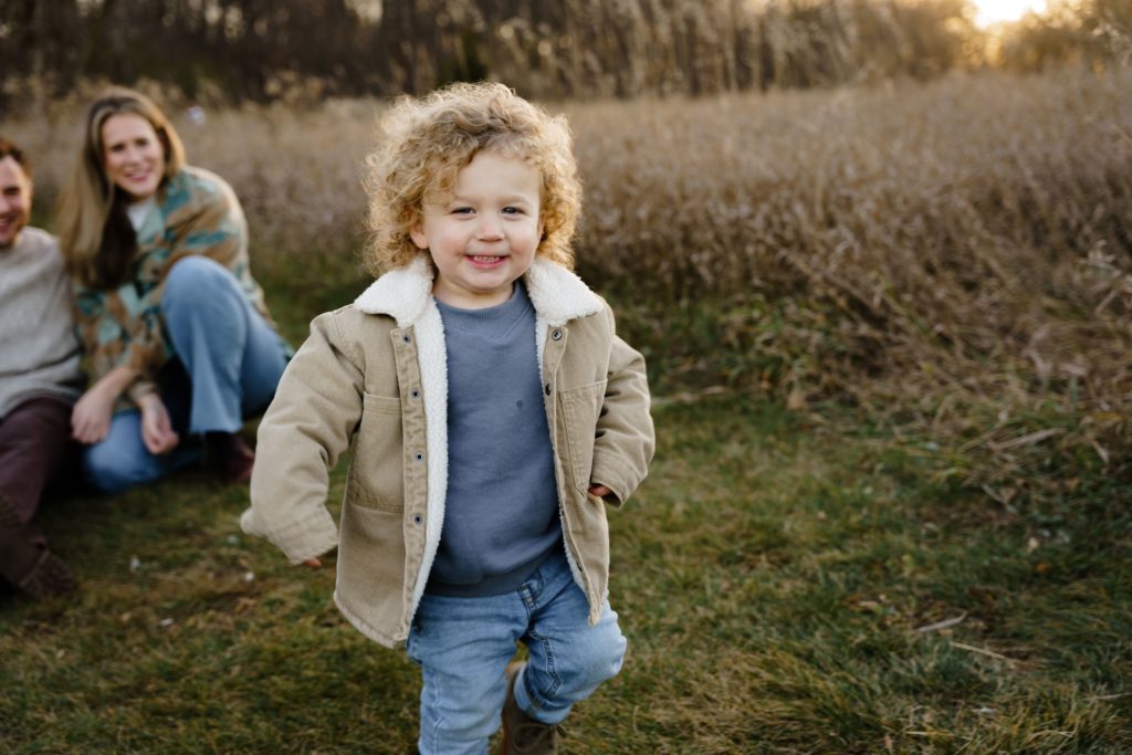 a boy laughs while running with his parents sitting in the background during their session with michigan family photographer