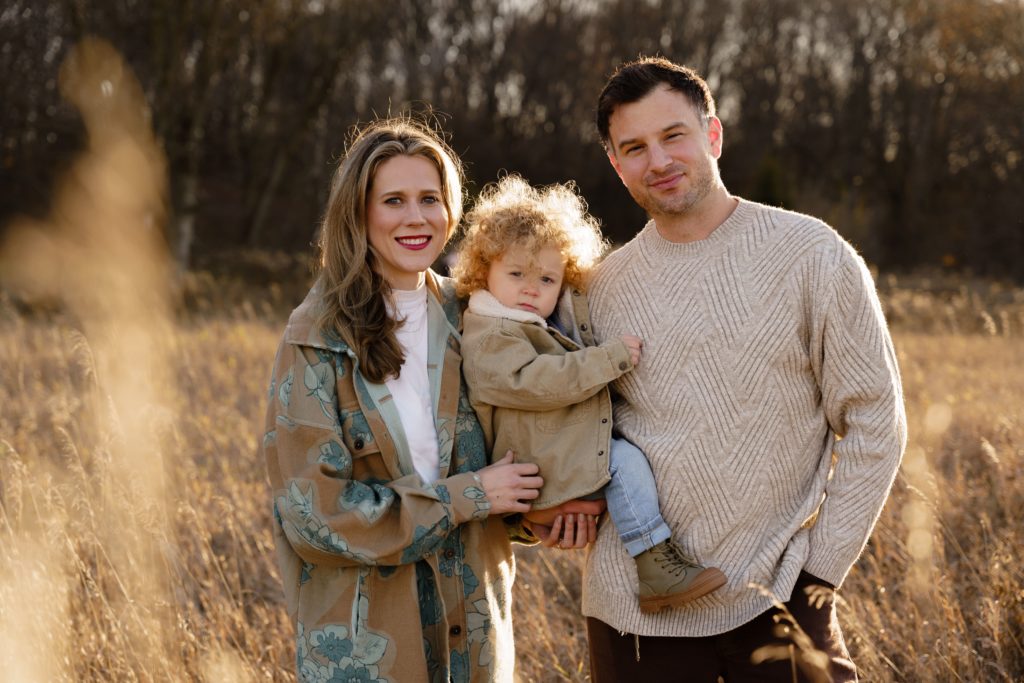 a family of three smile together for family portraits photography in an october field at golden hour