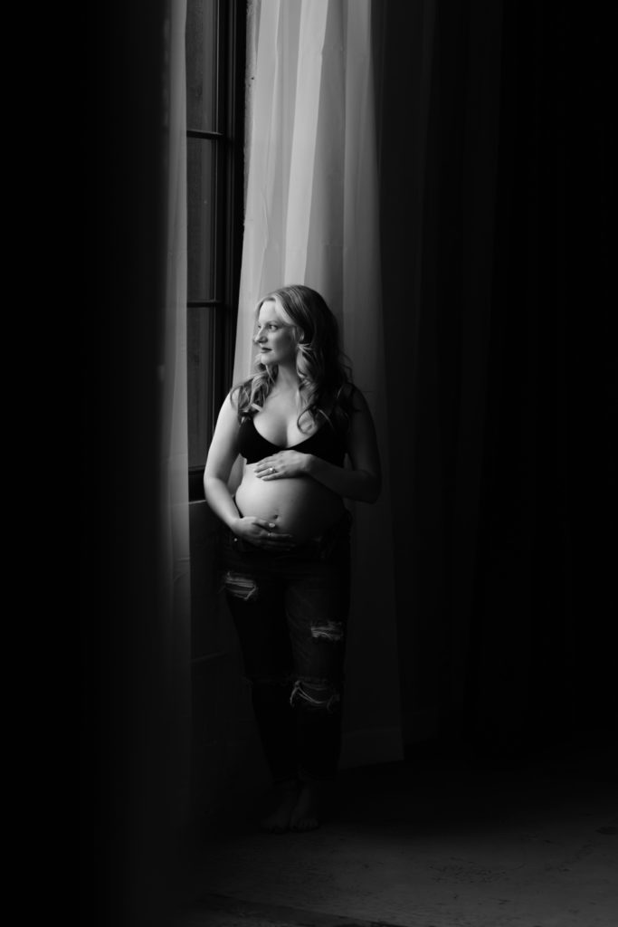 a filmy style portrait from a maternity photoshoot, chelsea stands by a window with light, sheer drapes