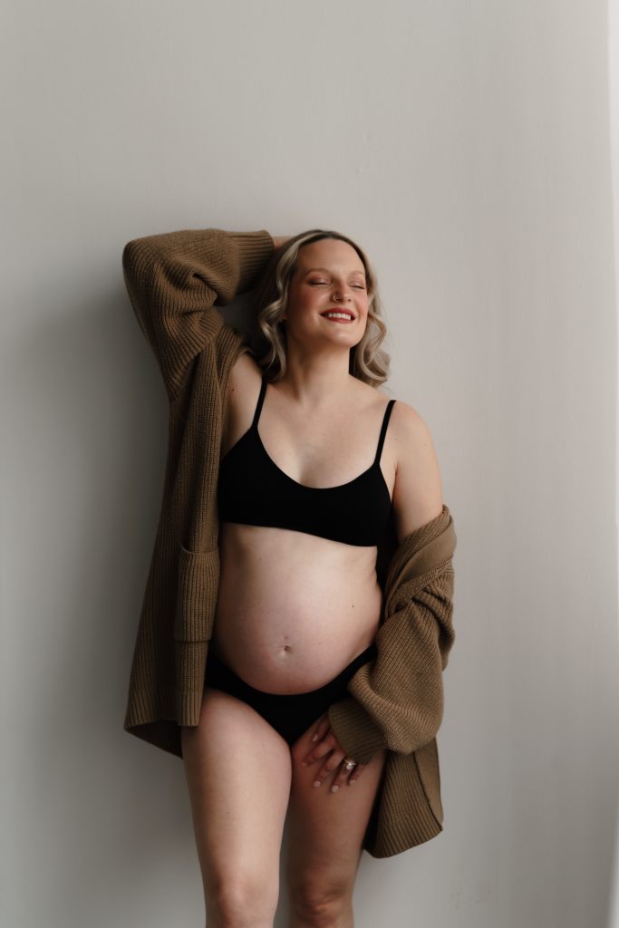 chelsea smiles while wearing a cardigan and lingerie set with an arm above her head during her maternity photoshoot