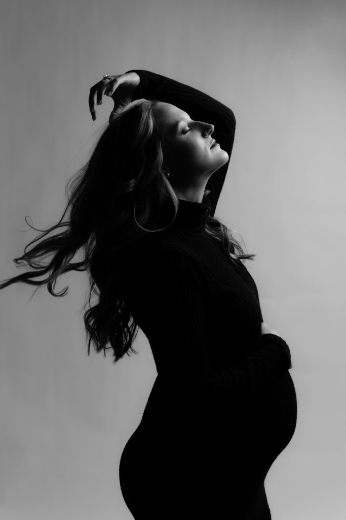 a woman basks in the studio light while swooshing her hair duirng her maternity photoshoot