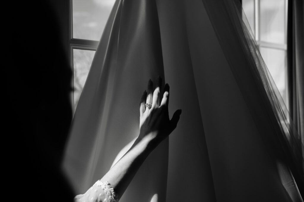 the bride reaches a hand out to her wedding dress hanging in front of a window