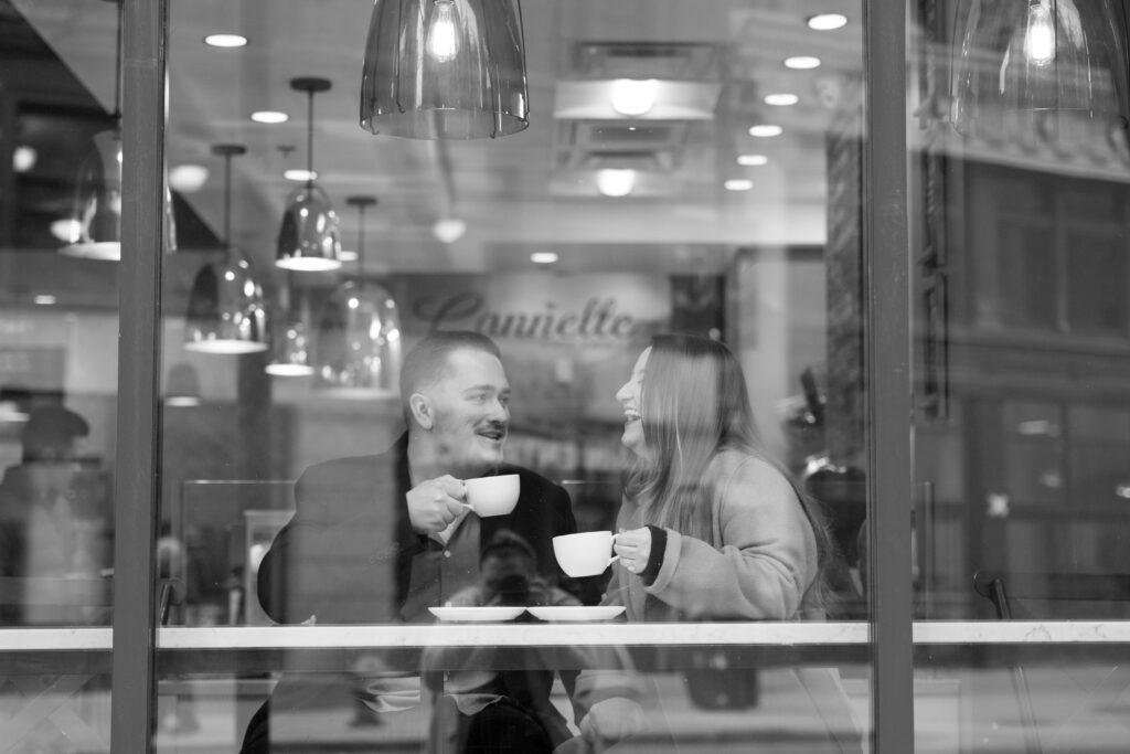 jane and john laugh over coffee on the opposite side of the window from their michigan engagement photographer