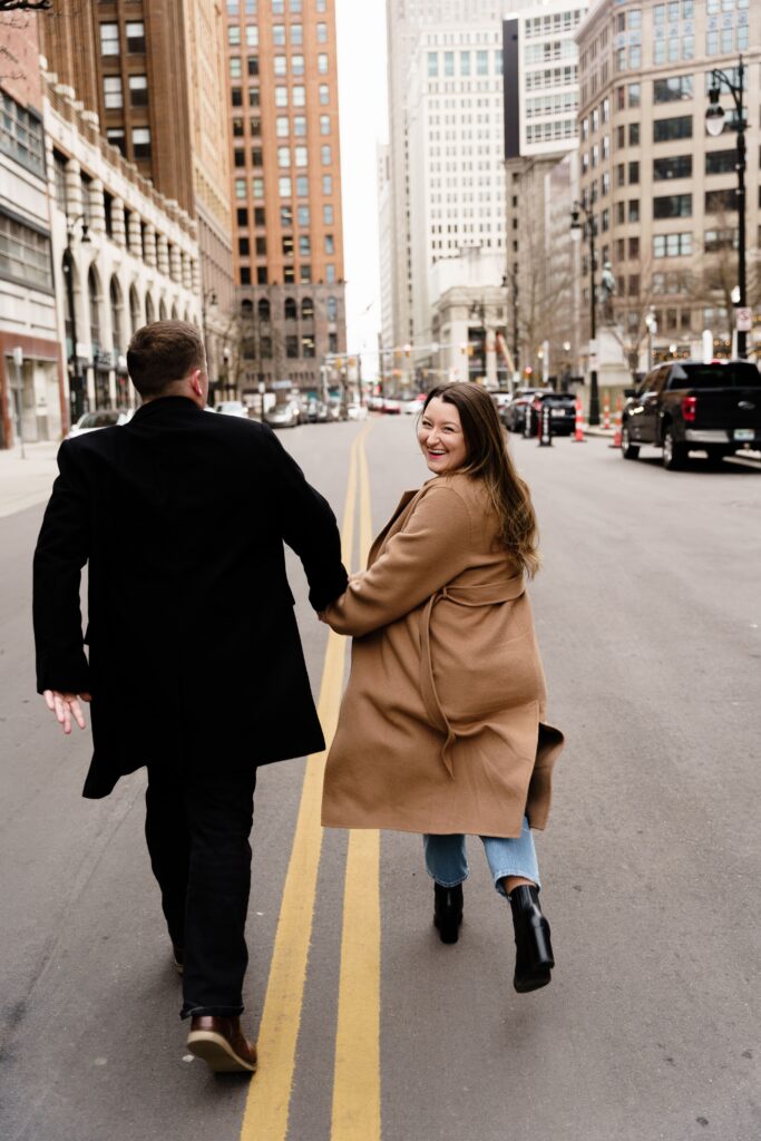 jane smiles over her shoulder as she and john run across the downtown detroit side street