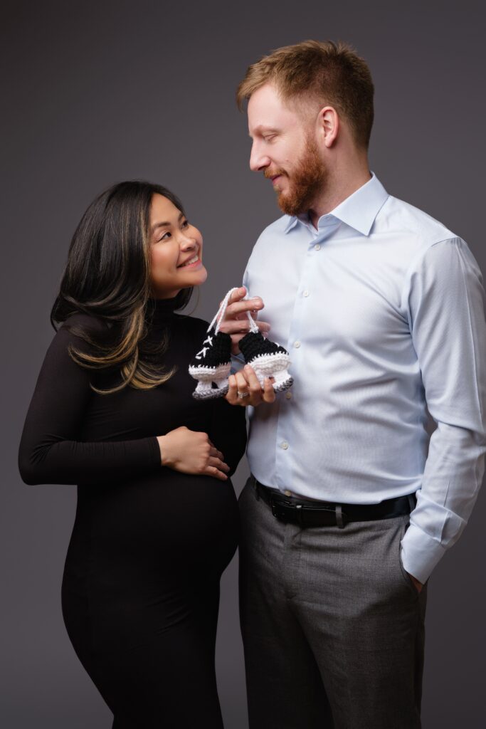 nat and andy smile as they hold up tiny crocheted ice skates during their lifestyle maternity photos