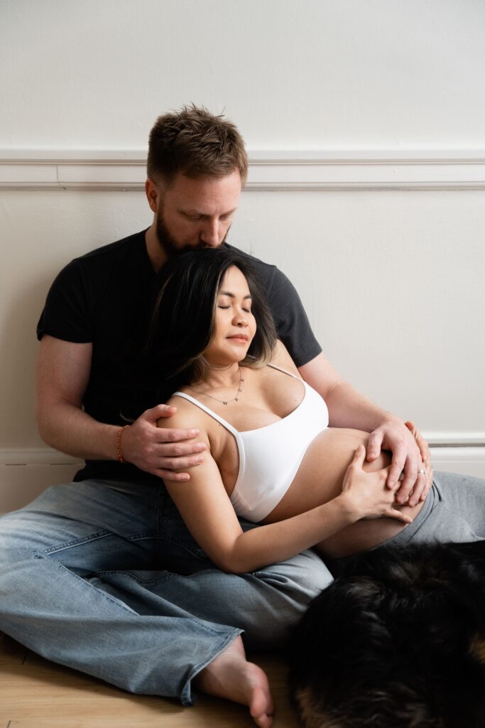 andy presses a kiss to nat's head as they sit on the floor with their pup during lifestyle maternity photos
