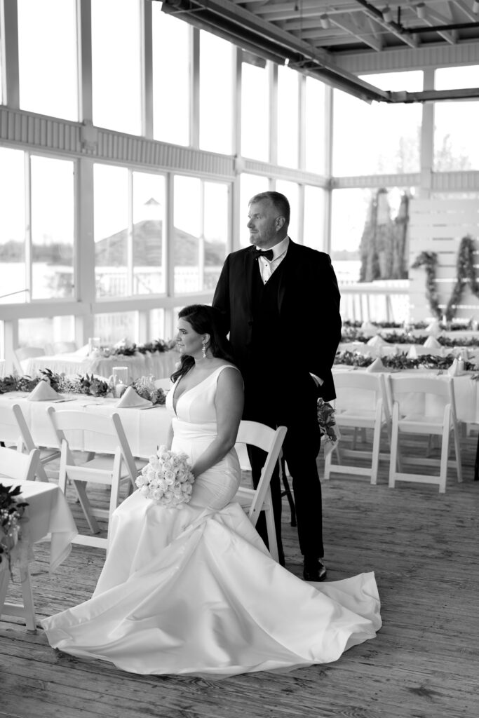 the bride sits in a chair with the groom standing behind her while posing for bridal portraits