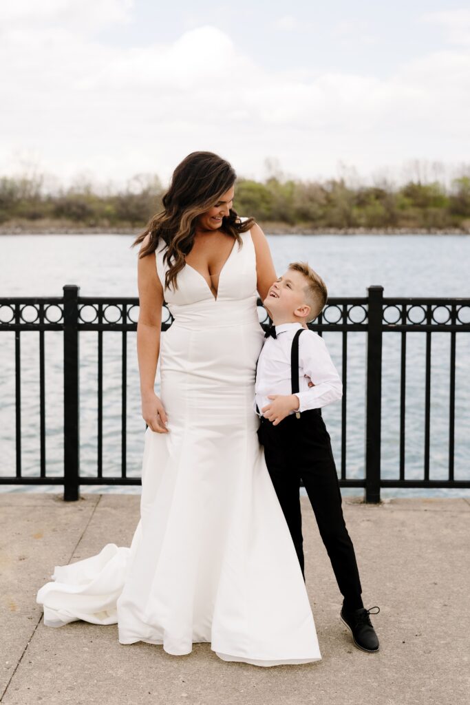 the bride smiles down at her son while standing waterfront and posing for bridal portraits